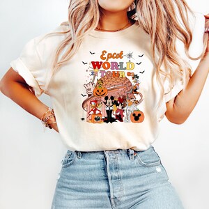 May include: Cream-colored t-shirt featuring a colorful graphic of Disney characters in Halloween costumes. The graphic includes the text "Epcot World Tour '82" with bats and pumpkins. The shirt has short sleeves and a crew neck.