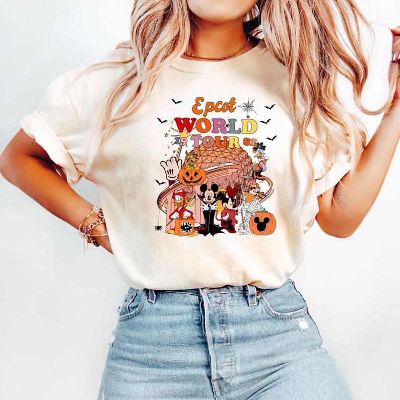 May include: Cream-colored t-shirt featuring a colorful graphic of Disney characters in Halloween costumes. The graphic includes the text "Epcot World Tour '82" with bats and pumpkins. The shirt has short sleeves and a crew neck.