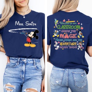 May include: A navy blue t-shirt with a Mickey Mouse design on the front and a Disney-themed classroom quote on the back. The quote reads "Welcome to our classroom where the magic never stops and the adventure never ends!"