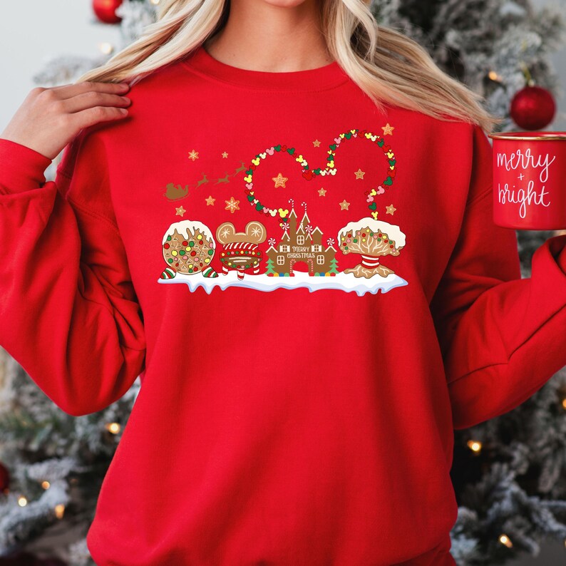 May include: Red Christmas sweatshirt featuring a gingerbread Mickey Mouse design. The design includes a gingerbread Mickey Mouse head with a string of lights and hearts around it. There are also gingerbread houses and trees on either side of the Mickey Mouse head. The text "Merry Christmas" is written on one of the gingerbread houses.