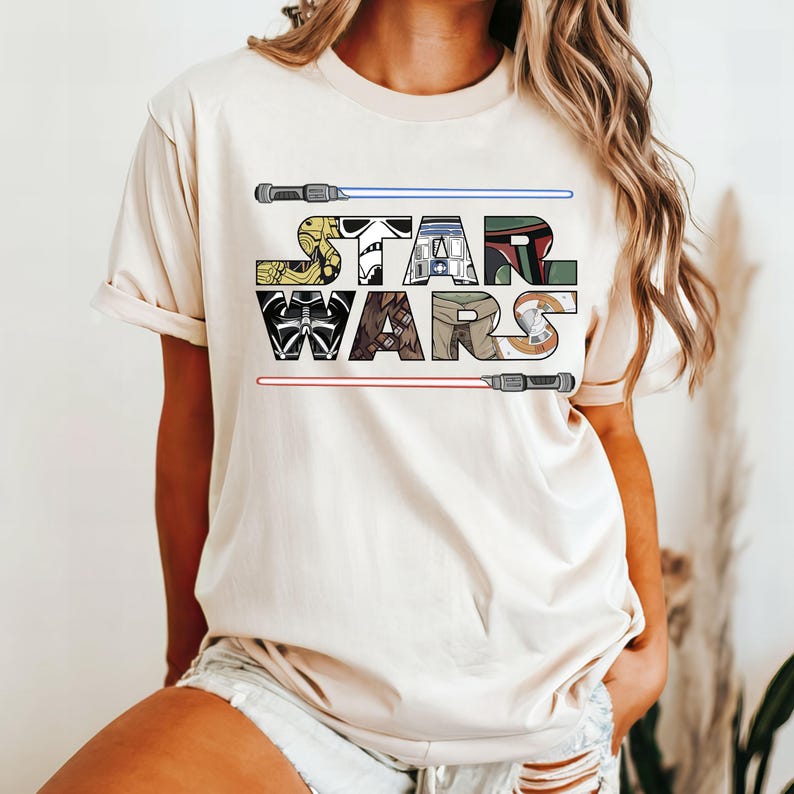 May include: A beige t-shirt with a graphic of the Star Wars logo. The logo is made up of images of characters from the Star Wars franchise, including Darth Vader, Chewbacca, R2-D2, and Yoda.