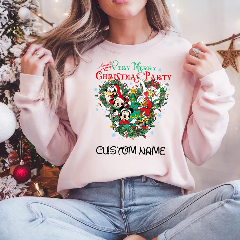 Może przedstawiać: R&oacute;żowa bluza z świątecznym motywem Myszki Miki z udziałem Myszki Miki, Minnie, Donalda, Goofy'ego i Pluto. Tekst na bluzie brzmi "Mickey's Very Merry Christmas Party" i "CUSTOM NAME".