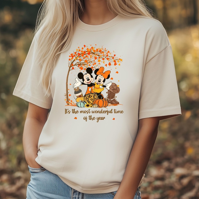 May include: Beige t-shirt with a fall-themed graphic featuring Mickey and Minnie Mouse. The design includes a tree with orange leaves, pumpkins, and the text "It's the most wonderful time of the year."