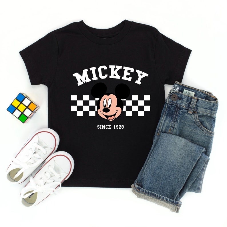 May include: Black t-shirt featuring Mickey Mouse with the word "MICKEY" in white block letters above a checkered pattern. Below the image is the text "SINCE 1928". The shirt is paired with blue jeans and white sneakers.