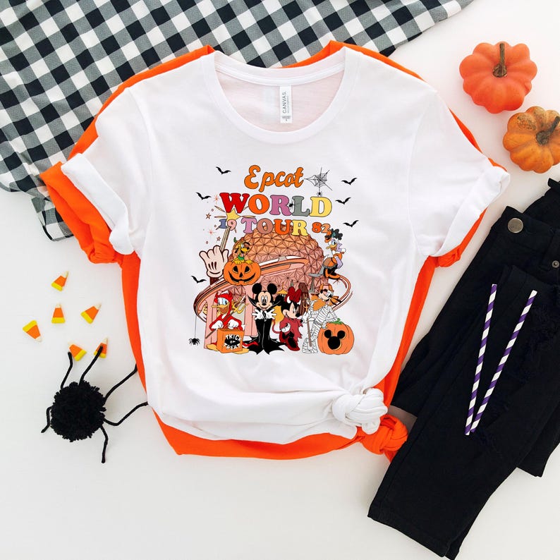 May include: White t-shirt with orange trim featuring a colorful graphic of Disney characters in Halloween costumes in front of a geometric structure. The text "Epcot World Tour" is above the graphic. Black pants and pumpkins are also in the image.