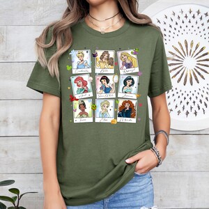 May include: Olive green t-shirt featuring nine cartoon portraits of Disney princesses in a grid layout. Each portrait is framed in a white border with the princess's name below. The princesses include Cinderella, Belle, Rapunzel, Ariel, Snow White, Jasmine, Tiana, Elsa, and Merida.