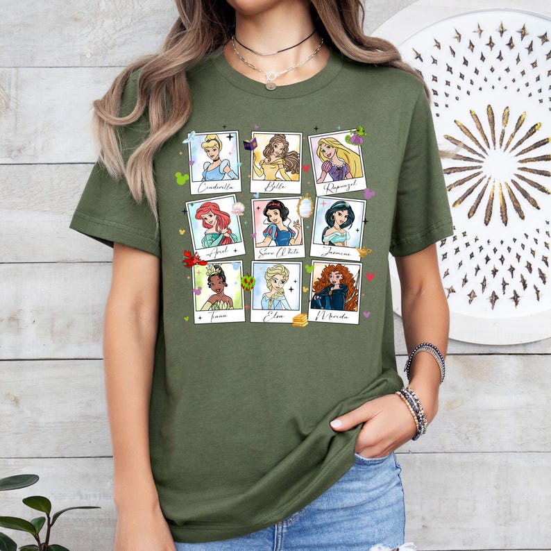 May include: Olive green t-shirt featuring nine cartoon portraits of Disney princesses in a grid layout. Each portrait is framed in a white border with the princess's name below. The princesses include Cinderella, Belle, Rapunzel, Ariel, Snow White, Jasmine, Tiana, Elsa, and Merida.