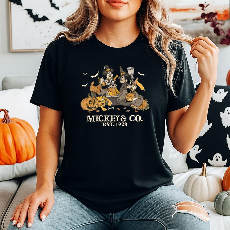 Pode incluir: T-shirt preto com um design de Halloween com Mickey, Minnie e Donald vestidos de bruxas, rodeados por ab&oacute;boras e morcegos. O texto "MICKEY & CO. EST. 1928" est&aacute; impresso abaixo.