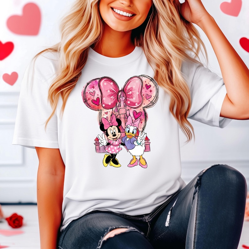 May include: White t-shirt featuring a graphic of Minnie Mouse and Daisy Duck with a pink bow and castle design. The cartoon characters are in a peace sign pose. Pink hearts are in the background.