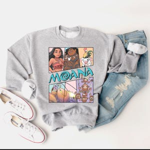 May include: A gray sweatshirt with a cartoon graphic of Moana and Maui from the Disney movie Moana. The graphic is in a comic book style with speech bubbles and a blue background. The text "Moana" is written in a blue font.