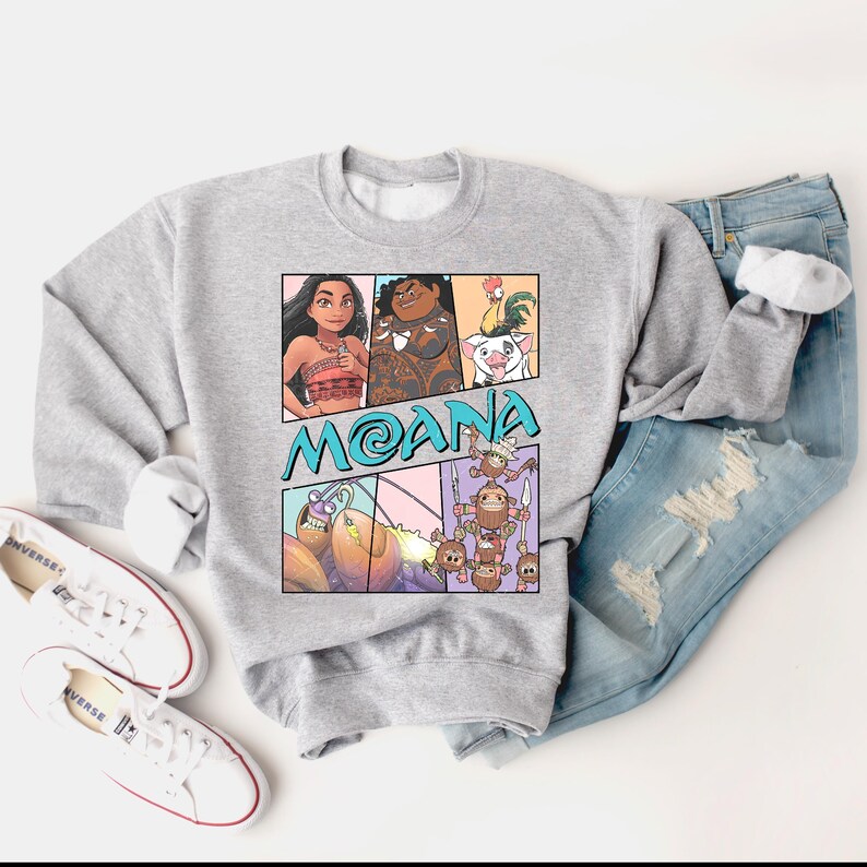 May include: A gray sweatshirt with a cartoon graphic of Moana and Maui from the Disney movie Moana. The graphic is in a comic book style with speech bubbles and a blue background. The text "Moana" is written in a blue font.