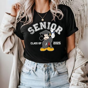 K&ouml;nnte beinhalten: Schwarzes T-Shirt mit einem wei&szlig;en Aufdruck von Mickey Mouse, der eine Graduation Cap tr&auml;gt und ein Diplom h&auml;lt. Der Text "Senior Class of 2025" ist auf dem Shirt aufgedruckt.