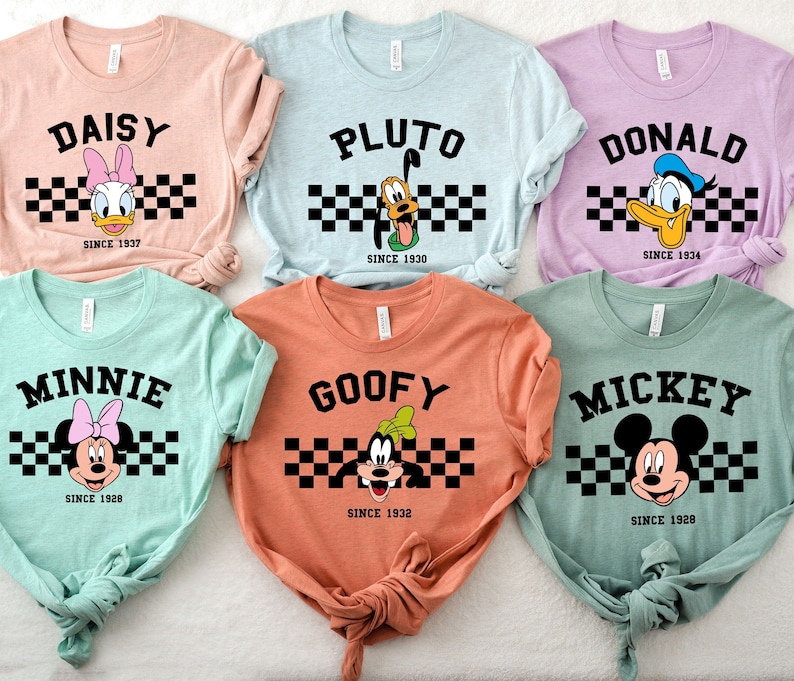 May include: Six colorful t-shirts featuring Disney characters: Daisy, Pluto, Donald, Minnie, Goofy, and Mickey. Each shirt has the character's name, a checkered stripe, and the year of their debut. The shirts are in various pastel colors.