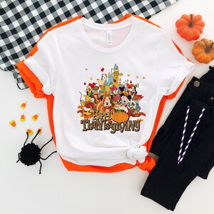 May include: White t-shirt with a colorful graphic featuring cartoon characters, a castle, and a pumpkin. The text "Happy Thanksgiving" is displayed below the graphic. The shirt is folded, with black pants and orange pumpkins in the background.