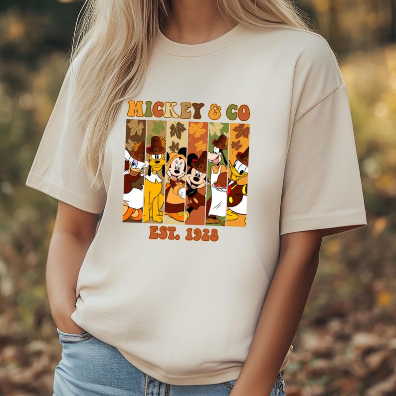 May include: Beige t-shirt featuring a colorful graphic of Disney characters in fall attire. The design includes the text "MICKEY & CO" and "EST. 1928" in a retro font. The characters are arranged in vertical stripes.