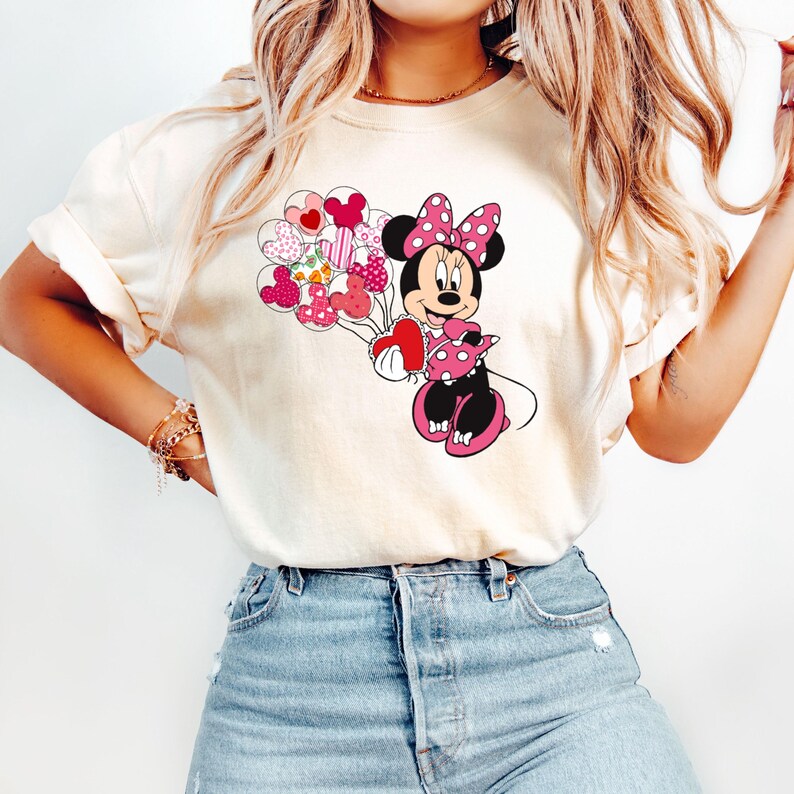May include: A white t-shirt with a graphic of Minnie Mouse holding a bunch of heart-shaped balloons. Minnie Mouse is wearing a pink bow and a red polka dot dress.