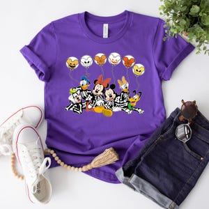 May include: Purple t-shirt with a graphic of Mickey Mouse, Minnie Mouse, Donald Duck, Goofy, and Pluto dressed as skeletons holding balloons with Halloween-themed faces.