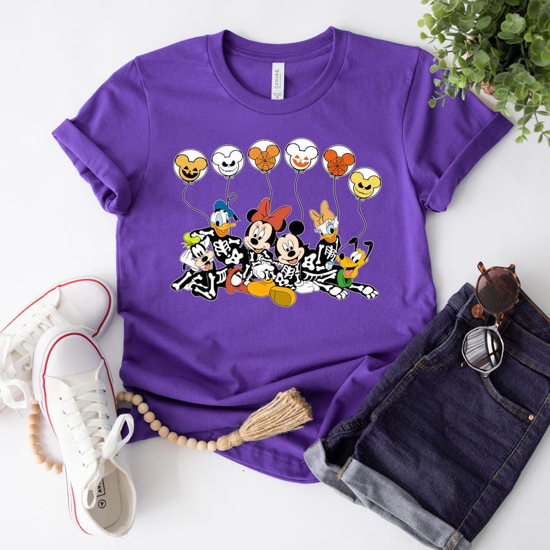 May include: Purple t-shirt with a graphic of Mickey Mouse, Minnie Mouse, Donald Duck, Goofy, and Pluto dressed as skeletons holding balloons with Halloween-themed faces.