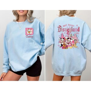 May include: Light blue sweatshirt with a custom design on the front and a Disneyland graphic on the back. The front features a pink framed letter "D" with a cartoon character and the word "Custom". The back has a Disneyland graphic with characters and text.