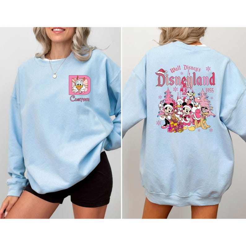 May include: Light blue sweatshirt with a custom design on the front and a Disneyland graphic on the back. The front features a pink framed letter "D" with a cartoon character and the word "Custom". The back has a Disneyland graphic with characters and text.