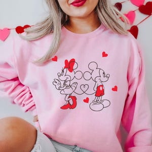 Op de afbeelding: Roze trui met een afbeelding van Mickey Mouse en Minnie Mouse die hand in hand staan. De afbeelding is in zwart en rood met hartjes eromheen.