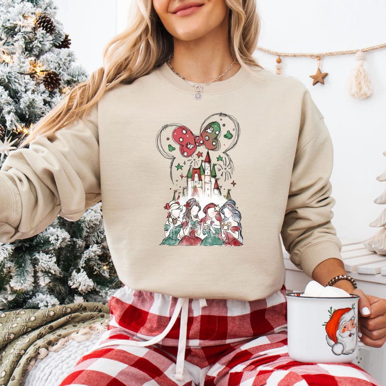 May include: Beige sweatshirt featuring a festive graphic. The design includes a red and green bow, a castle, and several figures. The sweatshirt is paired with red and white plaid pajama pants. A white mug with a Santa Claus design is also visible.