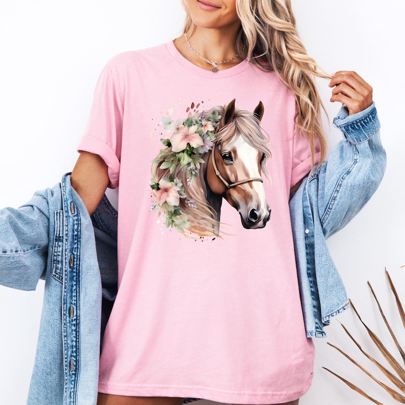 Peut inclure: T-shirt rose clair avec une illustration &agrave; l'aquarelle d'une t&ecirc;te de cheval orn&eacute;e de fleurs. Le cheval a une robe marron et blanche, un licol et une crini&egrave;re flottante. Le t-shirt est de couleur rose uni.