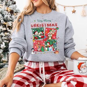 May include: A gray sweatshirt with a festive Christmas design. The design features four squares with Disney characters and the text "Very Merry Christmas" in red and green. The person is wearing red and white plaid pajama pants.