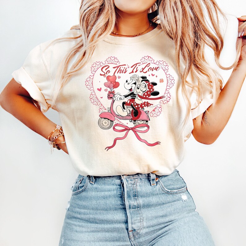 Puede incluir: Camiseta color crema con un dise&ntilde;o de Mickey y Minnie Mouse en una vespa rosa, rodeados por un dise&ntilde;o en forma de coraz&oacute;n con el texto "So This Is Love". El dise&ntilde;o incluye globos rosas y un gran lazo rosa.