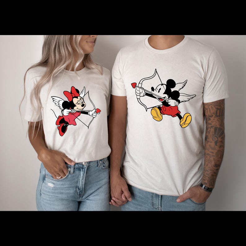 May include: A white t-shirt featuring a graphic of Mickey Mouse dressed as Cupid with wings and an arrow. The shirt is paired with a white t-shirt featuring a graphic of Minnie Mouse dressed as Cupid with wings and an arrow.