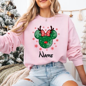 Może przedstawiać: R&oacute;żowy świąteczny sweter z zielonym i czerwonym wzorem głowy renifera Mickey Mouse i napisem "NAME" nadrukowanym poniżej.