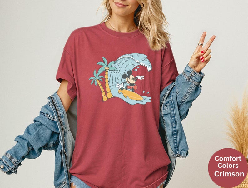 May include: Crimson red t-shirt featuring a graphic of Mickey Mouse surfing a wave. The design includes a palm tree and a yellow surfboard. The shirt is a Comfort Colors brand.
