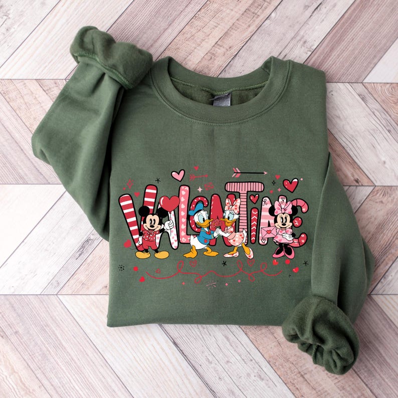 K&ouml;nnte beinhalten: Gr&uuml;ner Pullover mit einem Valentinstag-Design mit Mickey Mouse, Minnie Mouse, Donald Duck und Daisy Duck. Das Design enth&auml;lt das Wort "Valentine" mit Herzen und Pfeilen.