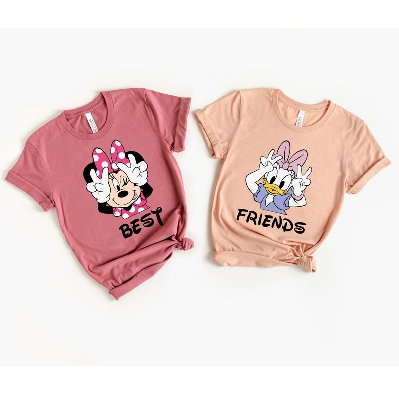 Może przedstawiać: Dwie koszulki w kolorze r&oacute;żowym i brzoskwiniowym z postaciami z kresk&oacute;wek. R&oacute;żowa koszulka przedstawia Minnie Mouse z napisem "BEST", a brzoskwiniowa koszulka przedstawia Daisy Duck z napisem "FRIENDS".