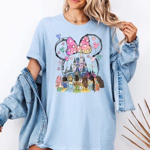 May include: Light blue t-shirt with a graphic of Cinderella, Snow White, Ariel, Belle, Rapunzel, and Tiana standing in front of Cinderella's castle with Mickey Mouse ears and a pink bow above the castle.