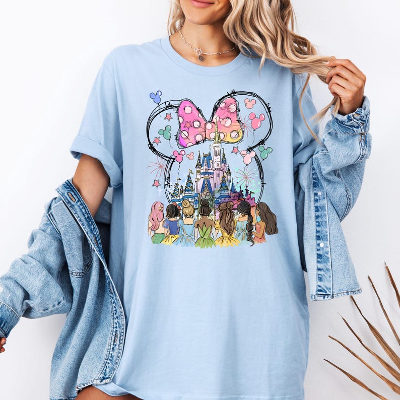 May include: Light blue t-shirt with a graphic of Cinderella, Snow White, Ariel, Belle, Rapunzel, and Tiana standing in front of Cinderella's castle with Mickey Mouse ears and a pink bow above the castle.