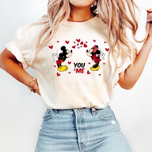 May include: White t-shirt with a graphic of Mickey Mouse and Minnie Mouse kissing. The graphic includes red hearts and the text "You & Me".