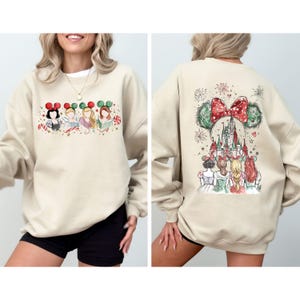 May include: Beige sweatshirt featuring a festive design. The front showcases cartoon characters with holiday-themed mouse ears and candy canes. The back displays a castle, fireworks, and characters with mouse ears, all in a watercolor style.