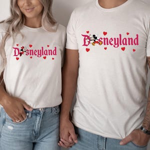 May include: A couple wearing white t-shirts with a pink Disneyland logo and Mickey Mouse and Minnie Mouse graphic. The shirts have red hearts around the logo.
