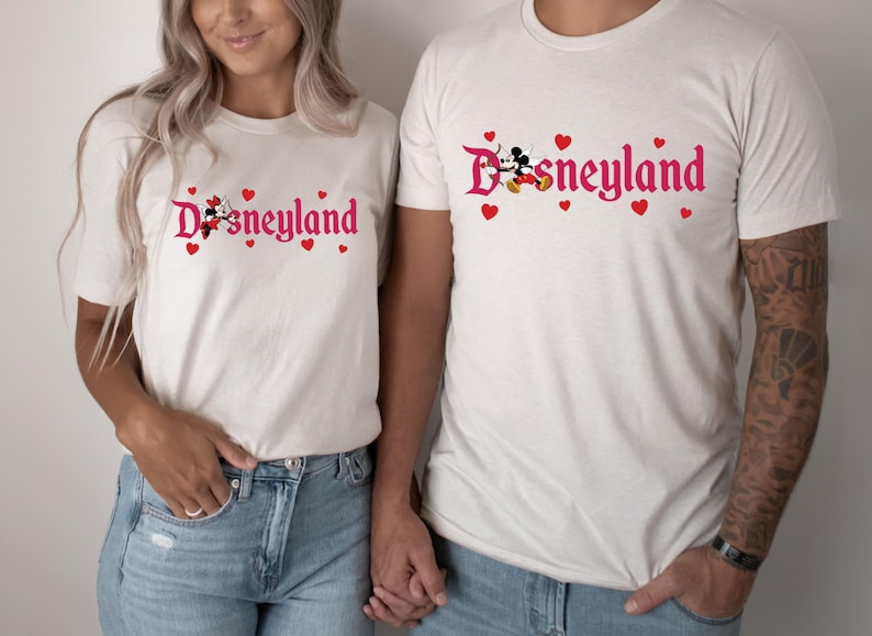 May include: A couple wearing white t-shirts with a pink Disneyland logo and Mickey Mouse and Minnie Mouse graphic. The shirts have red hearts around the logo.
