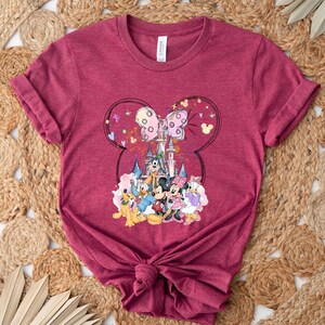 May include: A maroon t-shirt with a graphic print of Mickey Mouse and friends in front of Cinderella's Castle. The graphic is surrounded by a Mickey Mouse head outline with a pink bow.