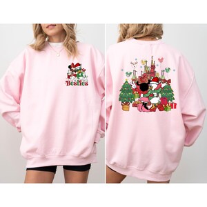 May include: A light pink sweatshirt featuring a festive graphic of Mickey and Minnie Mouse in Santa hats, with the word "Besties" in green and red. The back of the sweatshirt shows a Christmas scene with Donald Duck and other characters.