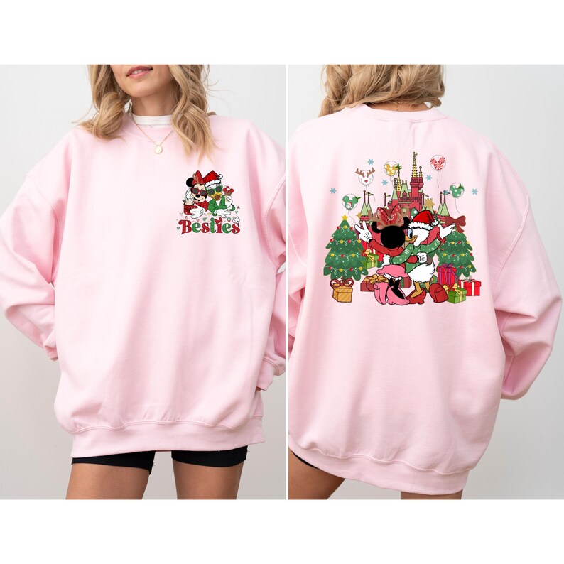 May include: A light pink sweatshirt featuring a festive graphic of Mickey and Minnie Mouse in Santa hats, with the word "Besties" in green and red. The back of the sweatshirt shows a Christmas scene with Donald Duck and other characters.