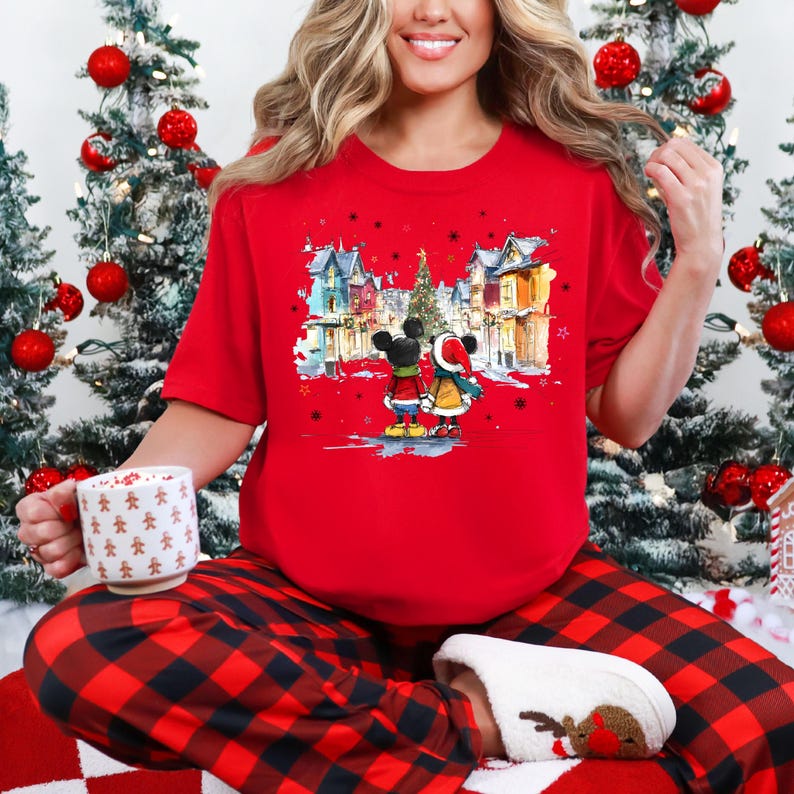 Op de afbeelding: Een rood T-shirt met een illustratie van Mickey en Minnie Mouse voor een winterlandschap. Het shirt wordt gedragen met een rood-zwarte geruite pyjamabroek. Een witte mok met peperkoekmannetjes is ook zichtbaar.