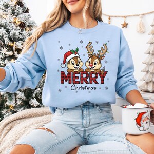 May include: Pale blue sweatshirt with a Christmas-themed graphic. It shows Chip and Dale characters, one in a Santa hat, the other with reindeer antlers, and the word "MERRY" in red plaid. Snowflakes and "Christmas" are also featured.