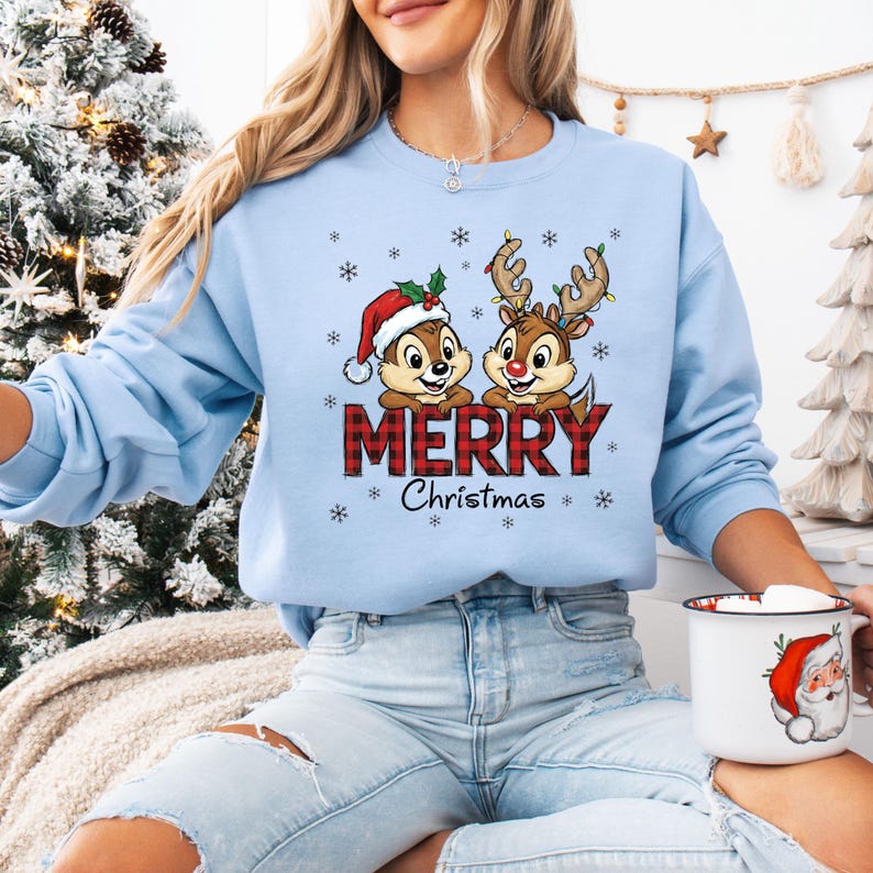 May include: Pale blue sweatshirt with a Christmas-themed graphic. It shows Chip and Dale characters, one in a Santa hat, the other with reindeer antlers, and the word "MERRY" in red plaid. Snowflakes and "Christmas" are also featured.