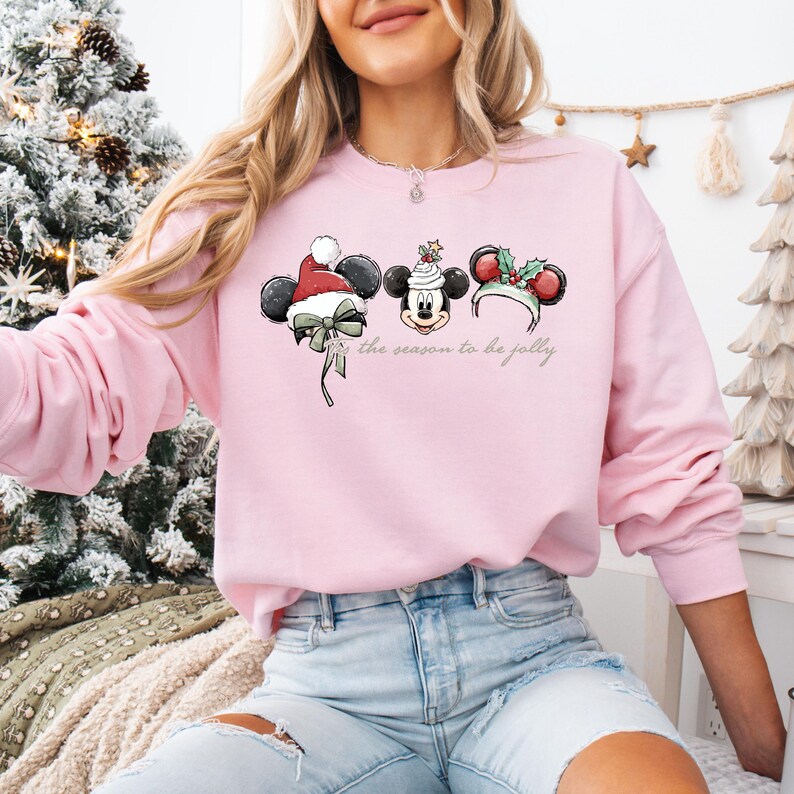 May include: A light pink sweatshirt featuring a festive Mickey Mouse design with holiday-themed ears. The design includes a Santa hat, a holly-adorned ear, and the phrase "the season to be jolly" in a script font. The sweatshirt is a long-sleeved crew neck.