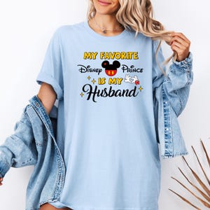 May include: Light blue t-shirt with the text "MY FAVORITE Disney Prince IS MY Husband" in black, yellow, and red. The design includes a Mickey Mouse head and a white glove. The shirt is worn with a denim jacket.