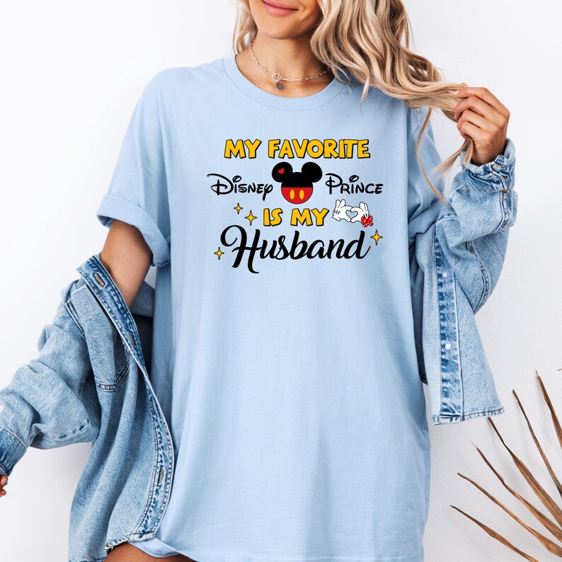 May include: Light blue t-shirt with the text "MY FAVORITE Disney Prince IS MY Husband" in black, yellow, and red. The design includes a Mickey Mouse head and a white glove. The shirt is worn with a denim jacket.