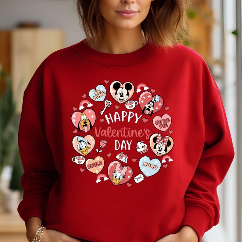 K&ouml;nnte beinhalten: Ein rotes Sweatshirt mit einem Valentinstags-Design. Das Design zeigt Herzen, Regenbogen und Disney-Figuren wie Mickey und Minnie Maus. Die Worte "Happy Valentine's Day" sind prominent dargestellt.